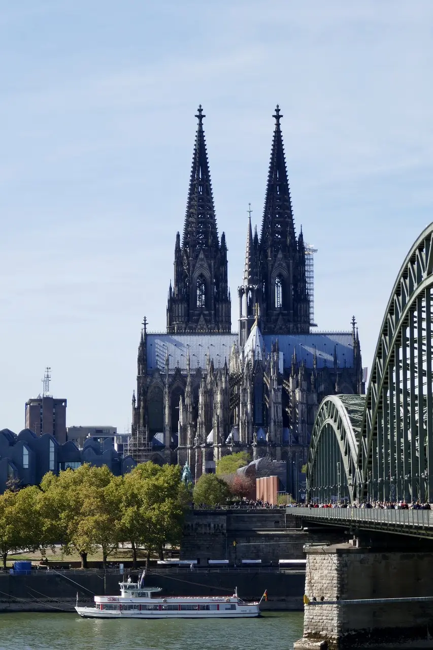 Kölner Dom mit Hohenzollernbrücke im Vordergrund, aufgenommen bei klarem Himmel vom Rheinufer aus.