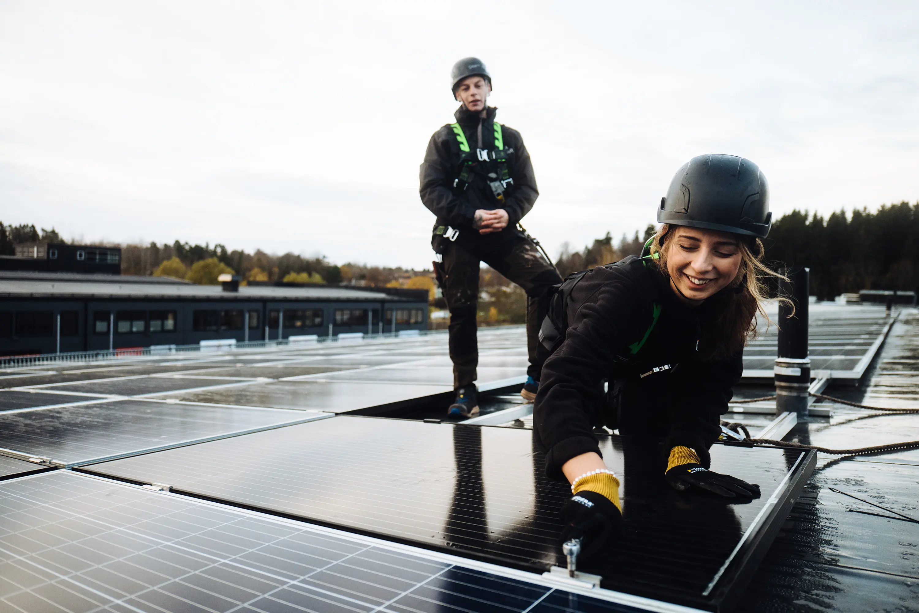 Zwei Monteur:innen installieren eine Solaranlage auf einem Flachdach, vorn kniet eine Frau und befestigt ein PV-Modul bei Tageslicht.