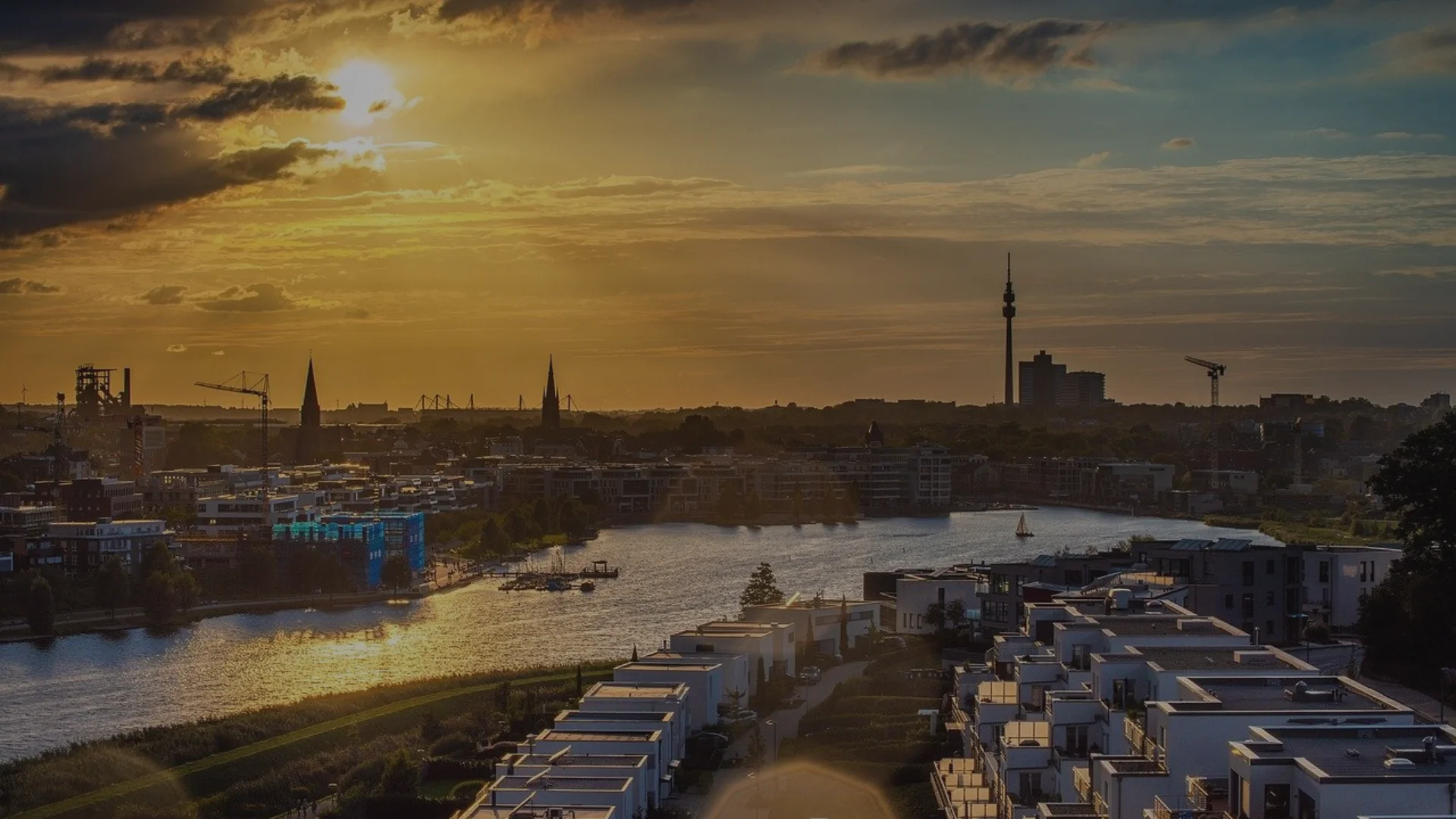 Stadtansicht mit Fluss bei Sonnenuntergang, im Vordergrund moderne Wohnhäuser, im Hintergrund der Dortmunder Fernsehturm und Industriebauten.