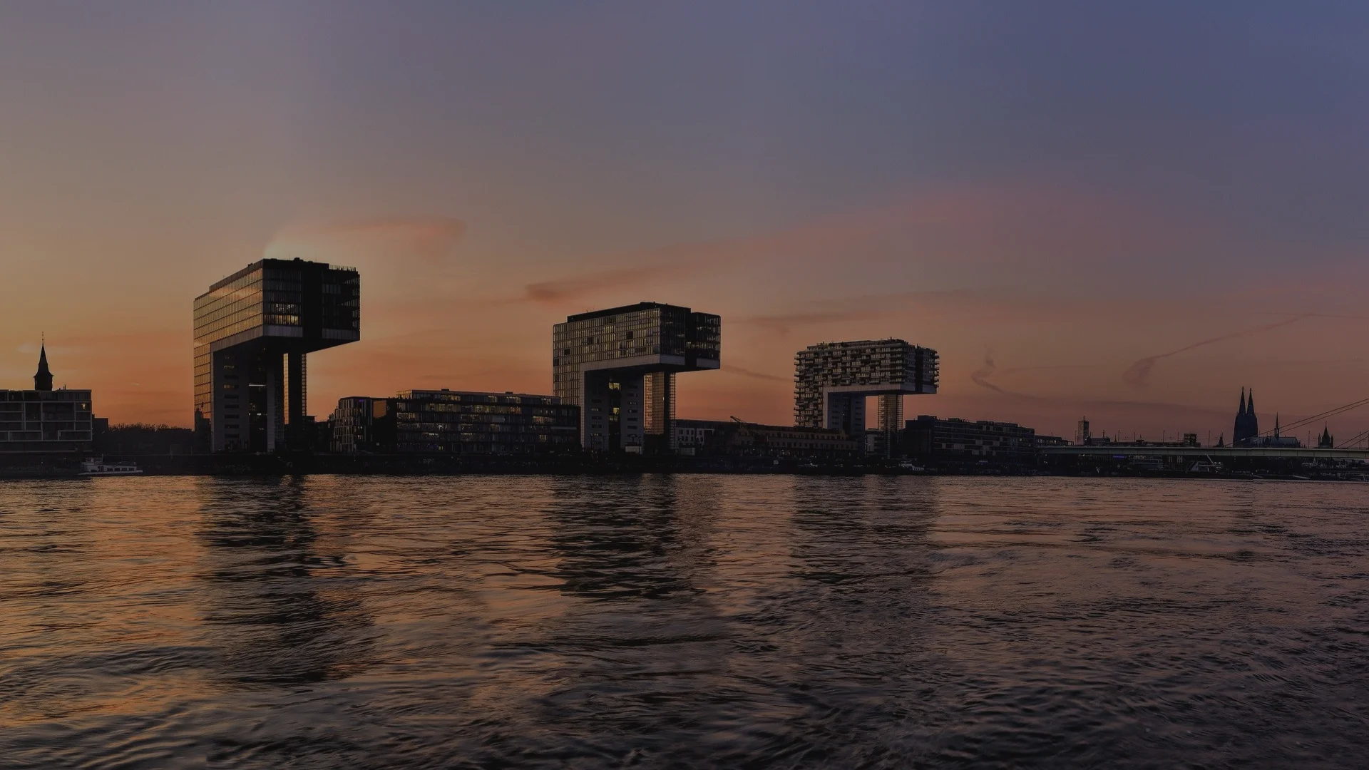 Blick auf die Kranhäuser am Rheinufer in Köln bei Sonnenuntergang, gespiegelt im ruhigen Wasser des Rheins.