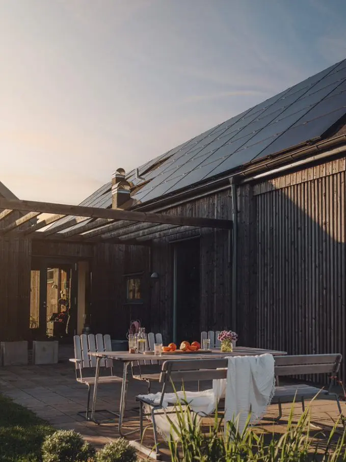 Terrasse mit gedecktem Tisch vor modernem Holzhaus mit großer Photovoltaikanlage auf dem Schrägdach im Abendlicht.